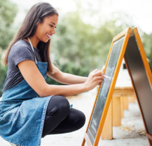 female barista writing menu on sidewalk chalkboard